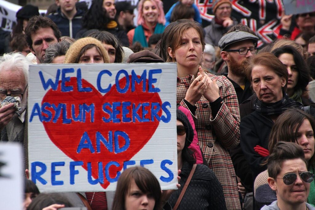 Photo of a rally with a woman holding a sign that reads, "welcome asylum seekers and refugees"